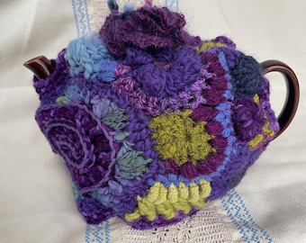 May include: A crocheted teapot cozy in shades of purple, blue, and green. The cozy is made up of various crocheted squares and flowers.