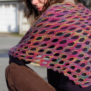 Puede incluir: Un chal calado multicolor en tonos rosa, morado, amarillo y verde. El accesorio de punto presenta un patrón ovalado repetitivo, creando un diseño delicado y aireado. El chal está drapeado sobre los hombros, mostrando su vibrante paleta de colores y textura.