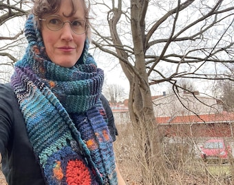 May include: A person wearing a blue, green, and orange crocheted scarf with a large circular design. The scarf is worn around the neck and head, and the person is standing in front of a tree.