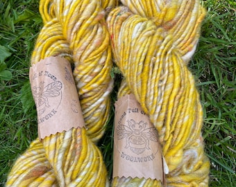 May include: Two skeins of yellow and white hand-dyed yarn with brown paper labels. The labels read "Head Full of Bees Woolwork" with a bee illustration. The yarn is thick and textured, resting on a bed of green grass.