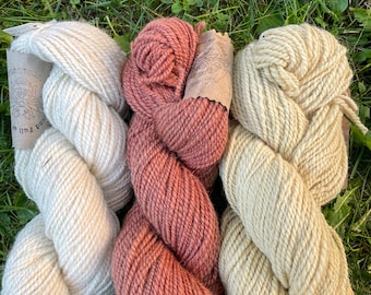 May include: Three skeins of yarn in cream, rose, and light yellow hues rest on a bed of green grass. The yarn is thick and appears to be hand-spun. The skeins are arranged side by side, showcasing their different colors and textures.