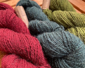 May include: Close-up of several skeins of yarn in shades of red, teal, and olive green. The yarn is thick and textured, with paper labels attached. The yarn is arranged on a wooden surface.