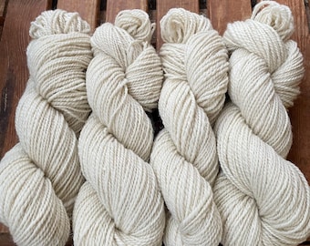May include: Four skeins of off-white yarn are arranged on a wooden surface. The yarn is thick and twisted, with a soft, textured appearance. The skeins are positioned close together, showcasing the natural color and fiber of the yarn. Ideal for knitting and crochet projects.