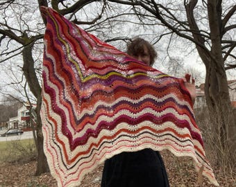 May include: A colorful crocheted shawl with a wavy pattern in shades of red, orange, pink, purple, and white. The shawl is held up by a person, with only their hand and arm visible.