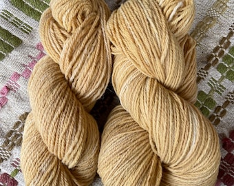 May include: Two skeins of golden yellow yarn with subtle white highlights, displayed on a patterned fabric with green, pink, and brown accents. The yarn appears soft and textured, suitable for knitting or crochet projects.