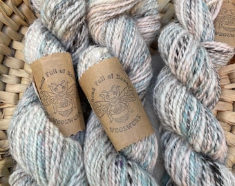 May include: Three skeins of speckled yarn in a woven basket. The yarn is a mix of gray, white, and pastel colors, with a label that reads "Head Full of Bees Woolwork" featuring a bee illustration. The yarn appears soft and textured.