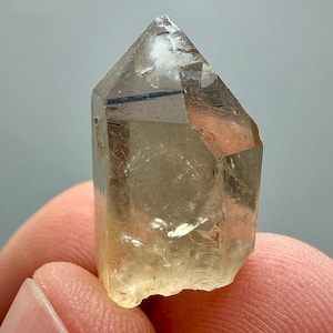Pode incluir: Um cristal claro e facetado com uma ponta. O cristal tem um tom castanho claro com inclusões escuras. O cristal é segurado entre dois dedos, mostrando sua forma natural e clareza. A superfície do cristal reflete a luz, destacando sua estrutura geométrica.