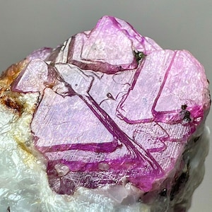 May include: Close-up of a raw, uncut gemstone. The crystal features a vibrant pink hue with a translucent quality, showcasing internal structures and lines. The stone is set against a white and brown background, highlighting its natural form.