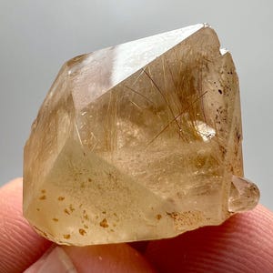 May include: A translucent, golden-brown quartz crystal with internal needle-like inclusions. The crystal has a faceted, geometric shape and a small, rounded protrusion at the base. The surface has a slightly rough texture with small, dark spots.