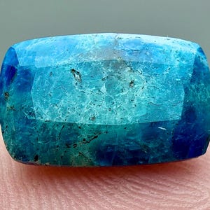 May include: A rectangular, faceted gemstone with a gradient of blue and teal hues. The gem's surface has a textured appearance, with darker blue areas at the edges and lighter teal in the center. The gem is resting on a pink surface.