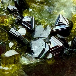 May include: Close-up of a mineral formation with dark, geometric crystals. The crystals have a reflective, metallic sheen, with a mix of dark and light bands. The surrounding matrix is a mix of green and yellow hues, creating a striking contrast.