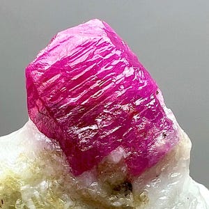 以下が含まれることがあります： 鮮やかなファセットルビーの結晶で、深いマゼンタ色を示しています。宝石は、その自然な形と質感を強調する、白と淡黄色の鉱物堆積物の背景に配置されています。ルビーの表面は光を反射し、その結晶構造を強調しています。