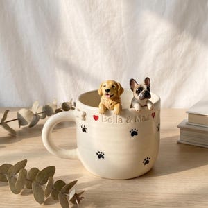 May include: A cream-colored ceramic mug featuring a sculpted golden retriever and a French bulldog peering over the rim. The mug is decorated with small black paw prints and red hearts, with the text Bella & Max embossed on the front surface.