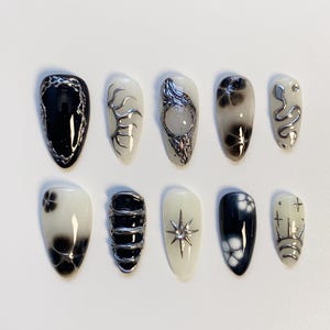 Puede incluir: Un conjunto de diez uñas artificiales con varios diseños. Las uñas presentan una mezcla de acentos negros, blancos y plateados. Los diseños incluyen patrones abstractos, motivos florales y adornos metálicos. Las uñas son de forma ovalada.