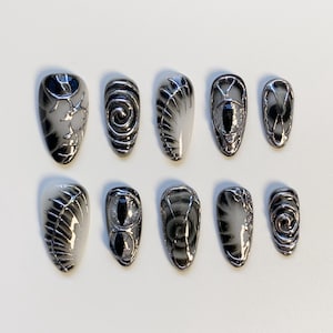 Puede incluir: Un conjunto de diez uñas artificiales con acabado brillante. Las uñas presentan una variedad de diseños, incluyendo espirales, telarañas y formas de ojo, todo en negro y plata. Las uñas están dispuestas sobre una superficie blanca.