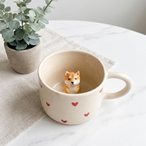 May include: Cream-colored ceramic mug with red heart accents. Inside the mug is a small, orange and white Shiba Inu dog figurine. The mug has a curved handle and is set on a white surface with a plant in the background.