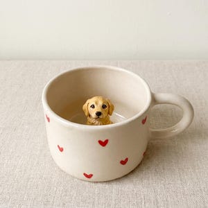 May include: Cream-colored ceramic mug with red heart accents. A small, detailed golden retriever puppy figurine sits inside the mug. The mug has a rounded handle and a smooth, matte finish.