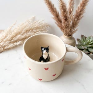 May include: Cream-colored ceramic mug with red heart accents. A small black and white cat figurine sits inside the mug. The mug has a curved handle and is on a white marble surface. Decorative dried plants are in the background.