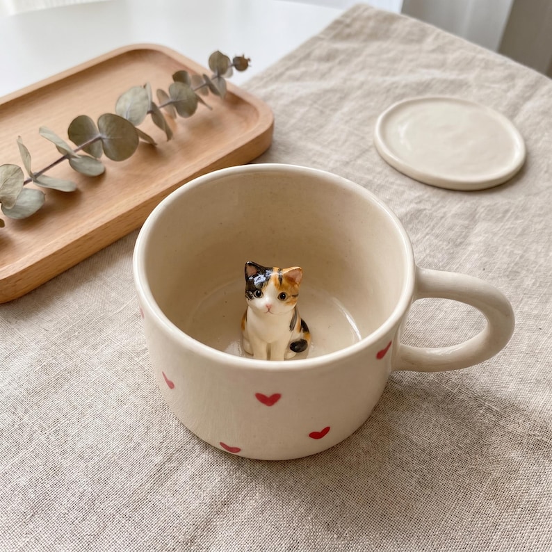 May include: Cream-colored ceramic mug with a small, three-colored cat figurine inside. The mug has a handle and is decorated with small red hearts. A matching saucer sits nearby, along with a wooden tray and eucalyptus sprig.