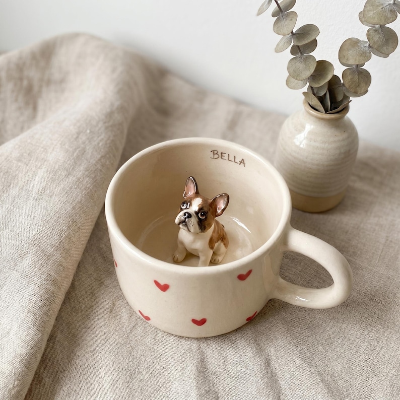 May include: Cream-colored ceramic mug with red heart accents and a small figurine of a French Bulldog inside. The word "BELLA" is written on the rim. A small vase with dried eucalyptus is in the background.