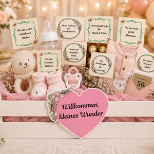 Puede incluir: Un set de regalo en caja de madera blanca que contiene un sonajero de cordero de peluche, un biberón, patucos rosas, un chupete, bombones y una prenda de bebé rosa. Incluye varias tarjetas con texto en alemán y un corazón rosa Willkommen, kleines Wunder.
