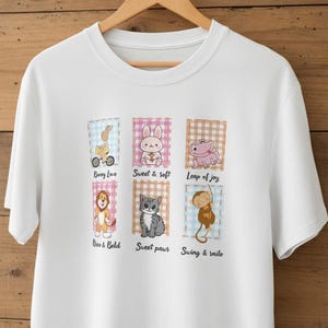 Könnte beinhalten: Weißes T-Shirt mit Rundhalsausschnitt und bunten Cartoon-Tiergrafiken. Das Design umfasst ein Häschen auf einem Roller, ein Kaninchen, einen Frosch, einen Löwen, eine Katze und einen Affen, jeweils mit einem Spruch. Das Shirt wird auf einem Holzbügel präsentiert.