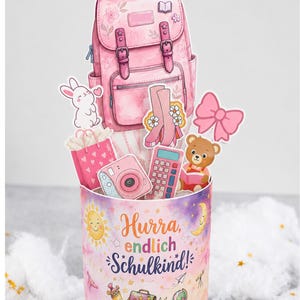 Puede incluir: Un recipiente cilíndrico con fondo degradado rosa y morado que presenta el texto Hurra, endlich Schulkind!, un sol, una luna y útiles escolares. Contiene recortes de una mochila rosa, un conejo, botas, un lazo, un oso y una cámara.