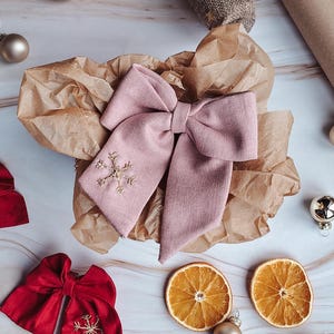 May include: A large, pale pink bow with a gold snowflake design, presented on brown paper. The image also features a red bow, dried orange slices, and gold baubles, creating a festive atmosphere.