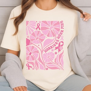 May include: Cream-colored t-shirt featuring a pink floral design with the words "Warriors Wear Pink." The design includes pink ribbons and flowers, promoting breast cancer awareness. The shirt is a casual wear item.