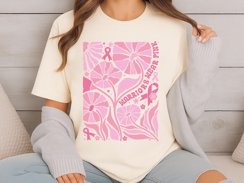 May include: Cream-colored t-shirt featuring a pink floral design with the words "Warriors Wear Pink." The design includes pink ribbons and flowers, promoting breast cancer awareness. The shirt is a casual wear item.
