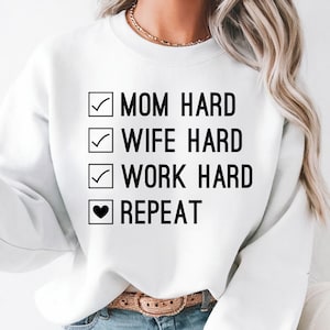 May include: White sweatshirt with the text "MOM HARD, WIFE HARD, WORK HARD, REPEAT" in black. Each line is preceded by a checkmark, except for "REPEAT", which is preceded by a heart symbol. The sweatshirt is worn with blue jeans and a brown belt.