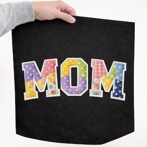 Może przedstawiać: Czarny materiał z napisem "MOM" w kolorowych literach patchworkowych. Litery są obrysowane na biało i mają różne wzory, w tym grochy, nadruki kwiatowe i kratę vichy. Ręka trzyma materiał, z kwiatami w tle.