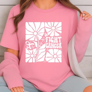 May include: Pink t-shirt with a white floral design and the words "FIGHT CANCER" in a square. The design includes a pink ribbon, a symbol of cancer awareness. The shirt is a short-sleeved crew neck.
