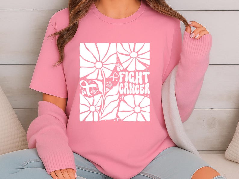 May include: Pink t-shirt with a white floral design and the words "FIGHT CANCER" in a square. The design includes a pink ribbon, a symbol of cancer awareness. The shirt is a short-sleeved crew neck.