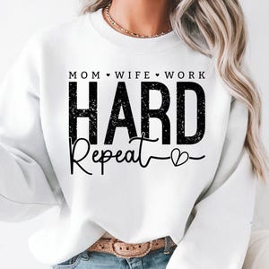 May include: White sweatshirt with the text "MOM WIFE WORK HARD Repeat" in black. The word "HARD" is in a bold, distressed font, and "Repeat" is in a cursive script. A heart symbol is at the end of the word "Repeat".