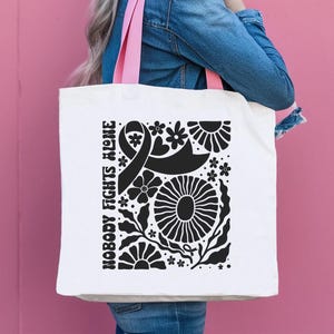May include: White tote bag with pink handles featuring a black floral design and the text "NOBODY FIGHTS ALONE." The design includes a ribbon, flowers, and leaves. The bag is held against a pink background.