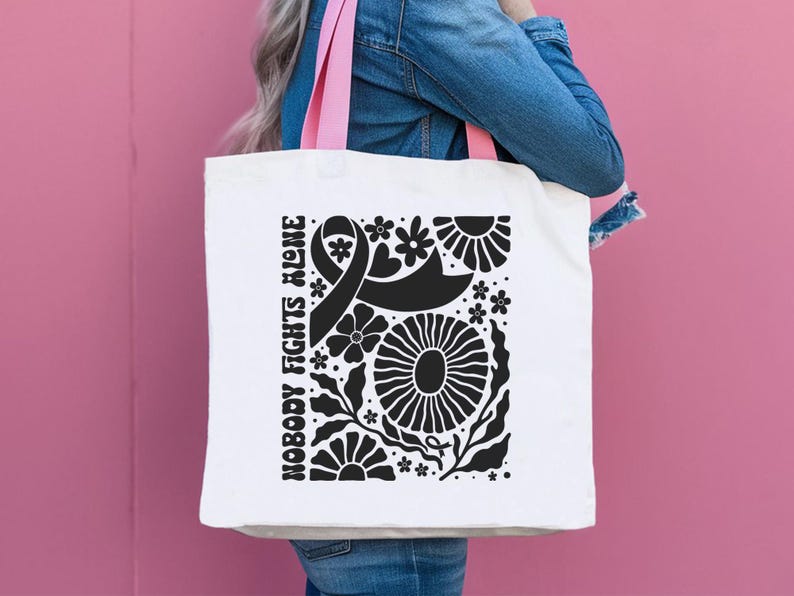 May include: White tote bag with pink handles featuring a black floral design and the text "NOBODY FIGHTS ALONE." The design includes a ribbon, flowers, and leaves. The bag is held against a pink background.