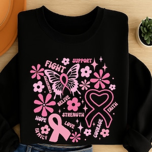 May include: Black sweatshirt with a pink breast cancer awareness design. The design includes a butterfly, flowers, ribbons, and words like "Fight", "Hope", and "Support". The sweatshirt is folded on a wooden surface.