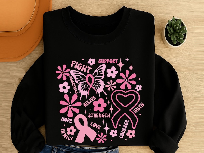 May include: Black sweatshirt with a pink breast cancer awareness design. The design includes a butterfly, flowers, ribbons, and words like "Fight", "Hope", and "Support". The sweatshirt is folded on a wooden surface.