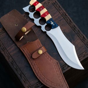 May include: A fixed-blade knife with a silver-toned steel blade and a handle featuring red, white, and blue stripes with a star emblem. The handle includes integrated finger holes. A brown leather sheath with brass snap closures is included.