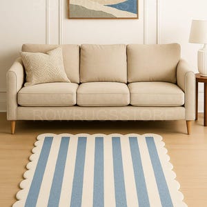 Op de afbeelding: Een beige bank met een kussen met patroon staat tegen een witte muur, boven een blauw-wit gestreept tapijt met een geschulpte rand. Een ingelijste landschapsschilderij hangt boven de bank.