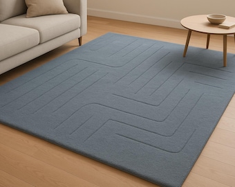 May include: A large, rectangular, blue rug with a geometric pattern of straight and curved lines. The rug is placed on a hardwood floor, with a beige sofa and a small wooden table with a bowl and book visible in the background.