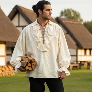 以下が含まれることがあります： クリーム色のルネサンスシャツ。フリルとレースアップフロントが特徴です。長袖シャツはゆったりとしたフィット感で、黒いパンツと茶色のブーツと組み合わせています。画像は屋外で、背景には藁葺き屋根の建物があります。