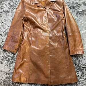 May include: A brown leather coat with a collared neckline and button closure. The coat has long sleeves and a tailored fit, with visible seams and a slightly textured surface. The coat is a medium length, reaching to the mid-thigh.