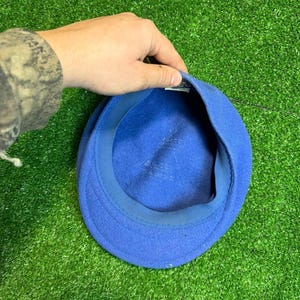 K&ouml;nnte beinhalten: Ein leuchtend blauer Hut aus Filz mit einer runden Krone und einem kleinen Schirm. Der Hut wird von einer Hand gehalten, wodurch das Innenfutter sichtbar wird. Der Hintergrund ist ein leuchtend gr&uuml;ner Kunstrasen.