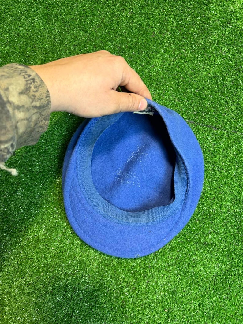 K&ouml;nnte beinhalten: Ein leuchtend blauer Hut aus Filz mit einer runden Krone und einem kleinen Schirm. Der Hut wird von einer Hand gehalten, wodurch das Innenfutter sichtbar wird. Der Hintergrund ist ein leuchtend gr&uuml;ner Kunstrasen.