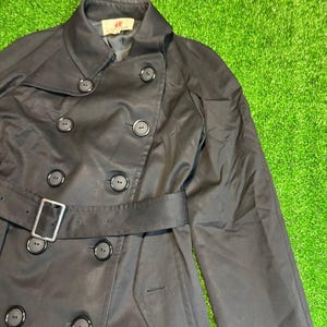 May include: A black trench coat with a double-breasted design, featuring large, round buttons and a matching belt with a silver buckle. The coat has a classic collar and a tailored fit.