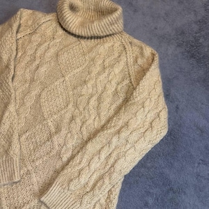 May include: A beige turtleneck sweater featuring a textured cable knit pattern throughout the body and sleeves. The garment has long sleeves with ribbed cuffs, a ribbed hem, and a thick, folded turtleneck collar made of a soft knit yarn.