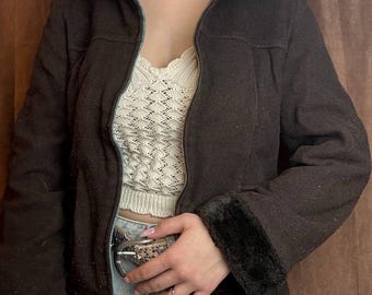 May include: A dark brown jacket with a hood and a zipper closure. The jacket has black faux fur trim on the cuffs and hood. The model is wearing a cream-colored top and light blue jeans.