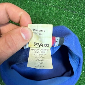 K&ouml;nnte beinhalten: Blauer Wollbarett mit einem Etikett mit der Aufschrift "CONTENTS 100% WOOL" und "MADE IN CUMBRIA ENGLAND BY TEAM 14 Stephen Nadia Gail Anna". Der Barett liegt auf einer gr&uuml;nen Oberfl&auml;che.